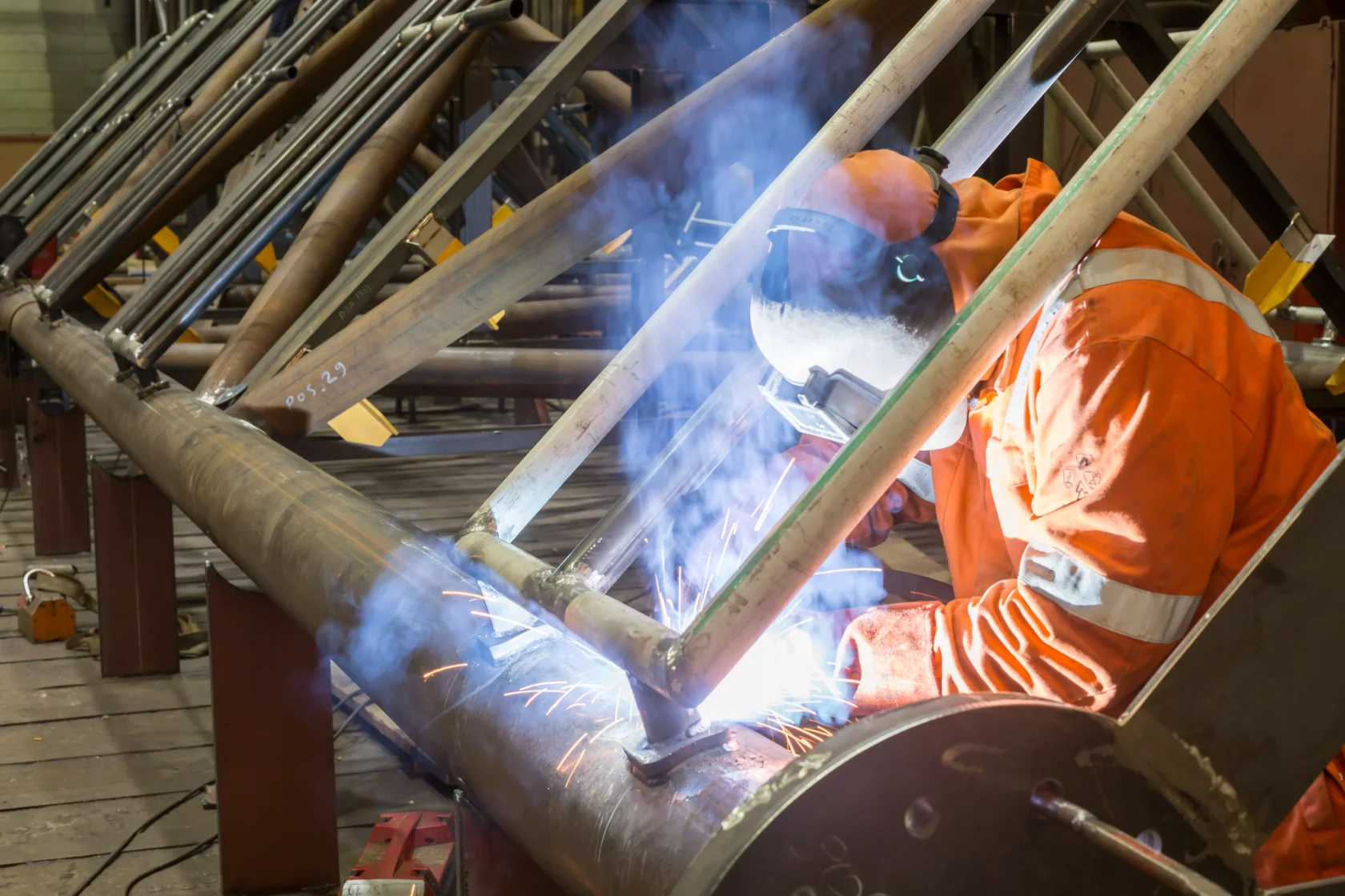 A welder in protective gear works on joining metal pipes in an industrial setting.