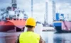 Engineer wearing a hard hat and safety vest, holding blueprints while inspecting ships at a shipyard.