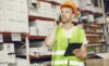 Worker in a safety vest and hard hat holding a clipboard while talking on the phone in a warehouse.