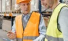 Two workers wearing safety vests and hard hats discussing in a warehouse.