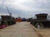 A shipyard with large industrial structures and cranes in the background, viewed from the deck of a vessel.