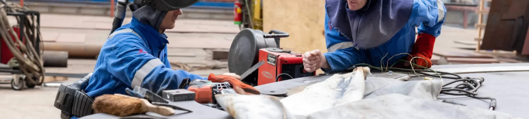 Two workers in protective gear, including welding helmets and gloves, engaged in welding and metalwork at an industrial site.