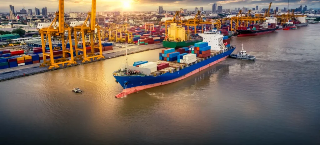 Container ship being guided by a tugboat into a busy port with colorful stacked containers and large cranes at sunset.