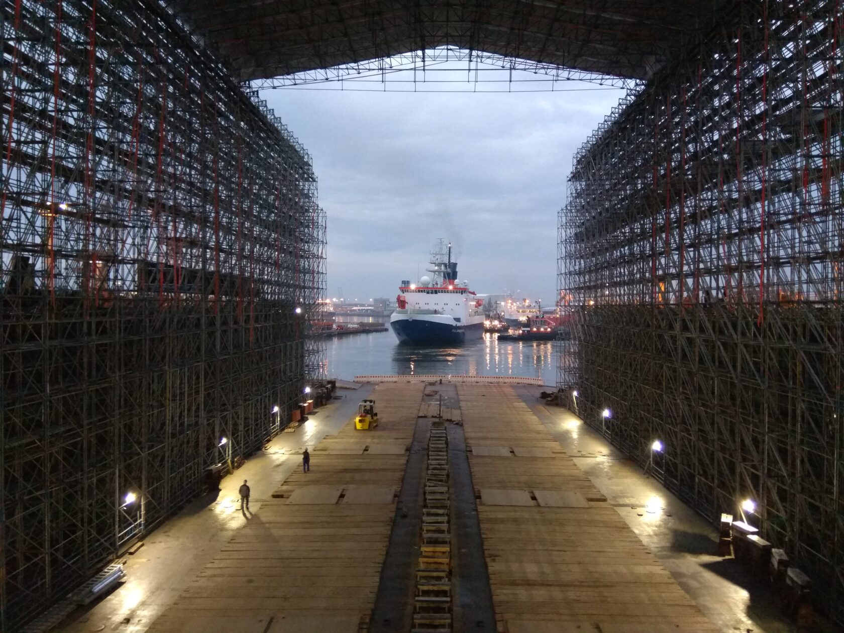 Large ship docked at a shipyard, viewed from inside a massive covered dry dock with scaffolding on both sides.